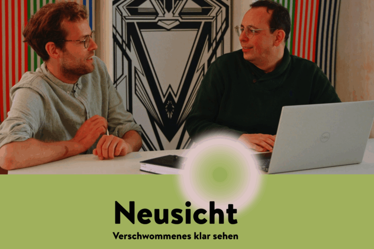 Neusicht - Verschwommenes klar sehen. Zwei Personen sitzen einander zugewandt an einem Schreibtisch und unterhalten sich. Vor ihnen ein geöffneter Laptop und ein Notizbuch.